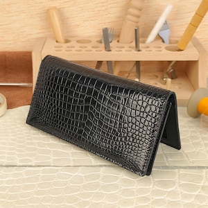 May include: A black leather wallet with a crocodile skin texture. The wallet is rectangular and has a folded design. The background includes wooden tools and a light-colored surface.
