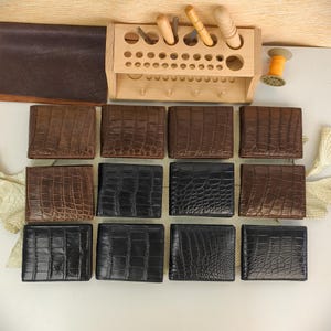Puede incluir: Una colecci&oacute;n de doce carteras plegables dispuestas en tres filas, con cuero grabado en relieve de cocodrilo en tonos marrones y negros. Sobre las carteras hay un organizador de herramientas de madera clara con varias herramientas.