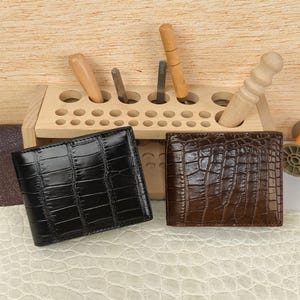 Puede incluir: Dos carteras plegables con una textura brillante de relieve de cocodrilo, una en negro y otra en marr&oacute;n oscuro. Est&aacute;n colocadas frente a un organizador de herramientas de madera clara que contiene varios utensilios de metal y madera.
