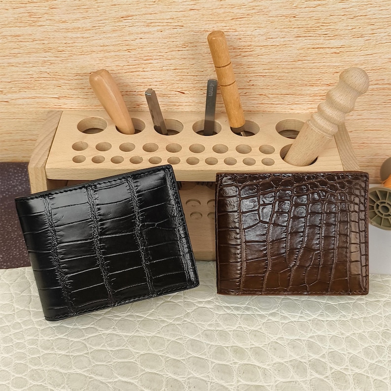 Puede incluir: Dos carteras plegables con una textura brillante de relieve de cocodrilo, una en negro y otra en marr&oacute;n oscuro. Est&aacute;n colocadas frente a un organizador de herramientas de madera clara que contiene varios utensilios de metal y madera.