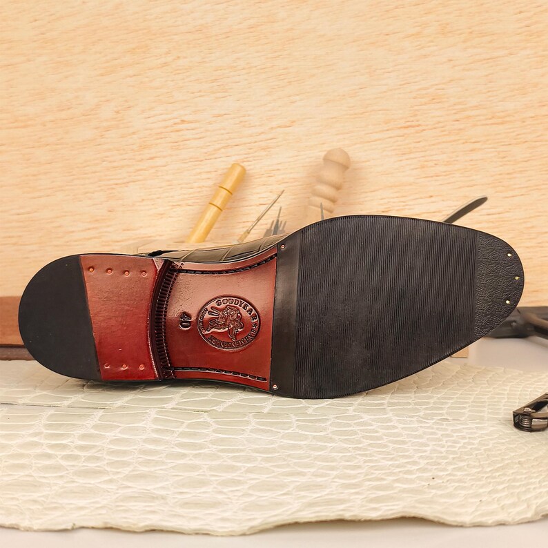 May include: Close-up of the sole of a black leather shoe with a Goodyear logo. The sole is black with a brown heel and a Goodyear logo. The shoe is on a white surface.