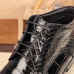 May include: Close-up of a black leather boot with a textured, crocodile-like pattern. The boot features black laces and a high-top design. The leather has a glossy finish, reflecting light. The overall style suggests a formal or dress shoe.