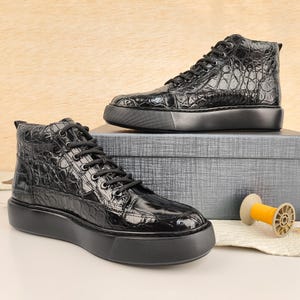 May include: Two black high-top sneakers with a textured, crocodile-like pattern. The sneakers have black laces and thick, black soles. One sneaker is positioned in front of the other, with a neutral background.