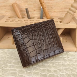 Puede incluir: Una cartera plegable de color marr&oacute;n oscuro con una textura brillante en relieve de piel de cocodrilo. La cartera tiene una forma rectangular con costuras visibles a lo largo de los bordes, mostrando un patr&oacute;n de escamas detallado en toda la superficie frontal.