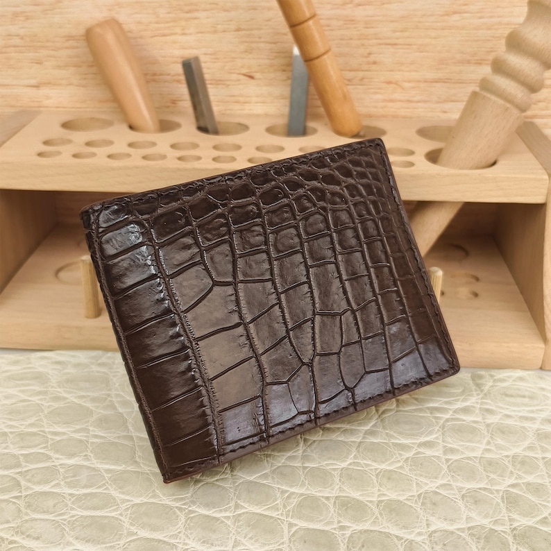 Puede incluir: Una cartera plegable de color marr&oacute;n oscuro con una textura brillante en relieve de piel de cocodrilo. La cartera tiene una forma rectangular con costuras visibles a lo largo de los bordes, mostrando un patr&oacute;n de escamas detallado en toda la superficie frontal.