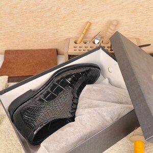 May include: A black leather shoe with a crocodile pattern sits inside a gray box. The shoe is shiny and appears to be new. The box is partially open, revealing white tissue paper. A wooden tool holder and leather pieces are in the background.