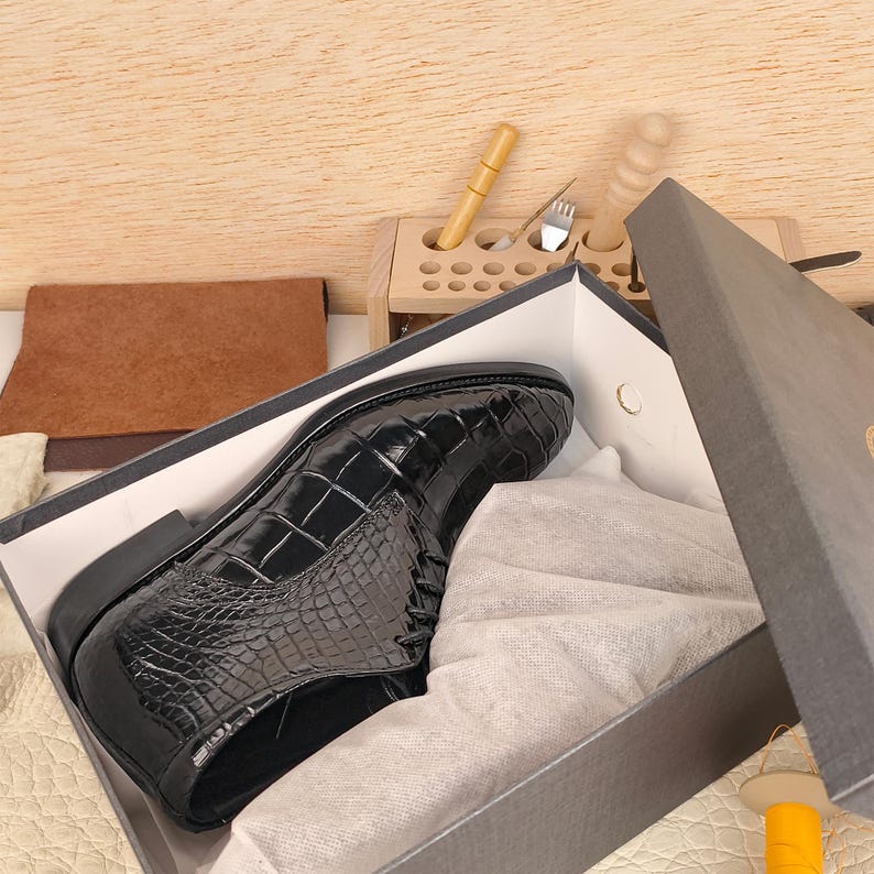 May include: A black leather shoe with a crocodile pattern sits inside a gray box. The shoe is shiny and appears to be new. The box is partially open, revealing white tissue paper. A wooden tool holder and leather pieces are in the background.