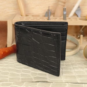 A sleek, handcrafted black genuine alligator leather bi-fold wallet with a glossy square-scale pattern, partially open to reveal internal card slots, displayed on a textured cream surface in an artisan workshop setting.