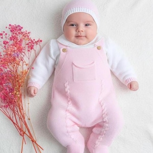 May include: A baby wearing a light pink knitted romper with a matching hat. The romper has a pocket and wooden buttons. The baby is lying on a white surface next to a bouquet of pink and orange flowers.