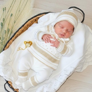 May include: A sleeping baby in a white and beige knitted outfit, including a hat, sweater, and trousers. The outfit features beige stripes and trim. A gold dummy with a chain rests on the blanket. The baby is in a woven basket.