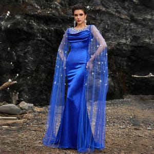 May include: A royal blue, floor-length gown with a fitted silhouette. The dress features a draped neckline and is paired with a sheer, cape-like overlay adorned with sparkling embellishments. The model is standing in front of a dark rock formation.