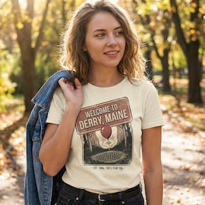 Puede incluir: Camiseta color crema con un gráfico de la película "It". El gráfico incluye un letrero que dice "Welcome to Derry, Maine" con un globo rojo y una imagen espeluznante. La persona lleva la camiseta en un parque con hojas de otoño.