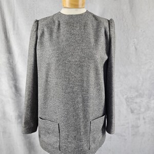 May include: A gray, long-sleeved top with a round neckline and two front pockets. The top is displayed on a mannequin. The fabric has a speckled texture. The sleeves have a slight puff at the shoulders. The hemline has a ribbed design.