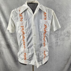May include: White short-sleeve shirt with orange floral embroidery. The shirt features a collared neckline, two chest pockets, and decorative vertical panels with orange floral designs. The shirt is buttoned down the front and has short sleeves.