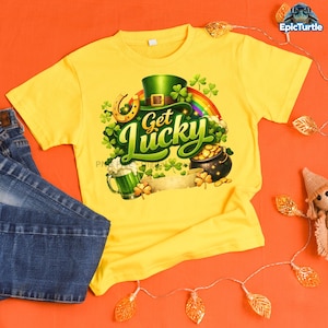 Puede incluir: Camiseta amarilla con un diseño del Día de San Patricio. El diseño incluye un sombrero verde, un arcoíris, una herradura, una olla de oro y las palabras "Get Lucky". También se muestra una jarra de cerveza verde y tréboles.