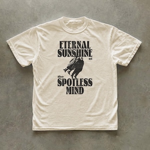 May include: An off-white short-sleeve t-shirt featuring a black graphic print on the chest. The design includes the text ETERNAL SUNSHINE of the SPOTLESS MIND with a central illustration of two figures embracing in a dark silhouette.