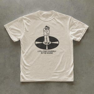 Op de afbeelding: Een gebroken wit T-shirt met korte mouwen en een zwarte afbeelding van twee figuren die op een vinylplaat staan. Onder de afbeelding staat de tekst: I STILL FIND YOURSELF IN THE SOUND. Het T-shirt heeft een ronde hals en een standaard pasvorm.