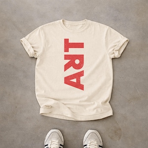 May include: A cream-colored short-sleeve t-shirt featuring the word ART printed vertically in a distressed red font. The shirt has a crew neckline and rolled sleeves, displayed with a pair of light-colored sneakers visible at the bottom edge.