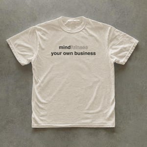 May include: A short-sleeved, off-white crew neck t-shirt featuring the black text "mindfulness" on the top line and "your own business" on the bottom line, centered on the chest. The shirt has a standard fit with a ribbed collar and hemmed sleeves.