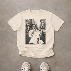 May include: A cream-coloured short-sleeve t-shirt featuring a black and white graphic print of a person in a chef's coat standing on a city street. The word NOMAD is printed in white capital letters across the centre of the graphic design.