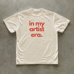 May include: A short-sleeved crew neck t-shirt in an off-white color. The front of the shirt features the text "in my artist era." printed in a bold, red, sans-serif font centered on the chest area of the garment.
