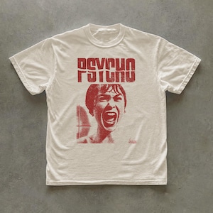 Pode incluir: Uma t-shirt de manga curta cor de creme com uma impressão gráfica vermelha. O design inclui a palavra PSYCHO numa fonte desgastada sobre uma ilustração pontilhada de uma pessoa a gritar com a boca bem aberta num cenário de duche.