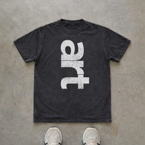 May include: A charcoal gray short-sleeve t-shirt featuring the word "art" printed vertically in large, distressed white capital letters centered on the chest. The garment has a crew neckline and a relaxed fit with a faded, vintage-style texture.