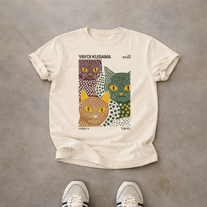 May include: A cream-colored short-sleeve t-shirt featuring a graphic print of three stylized cats covered in polka dots. The text reads YAYOI KUSAMA, 1998, TOKYO, and Japanese characters. The cats are colored in dark purple, green, and tan with yellow eyes.