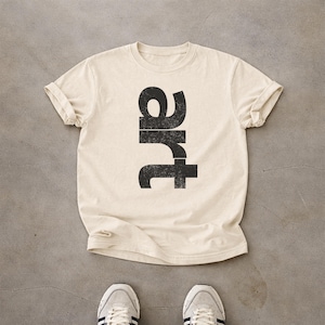 Peut inclure: Un t-shirt à manches courtes de couleur crème avec le mot "art" imprimé verticalement dans une police noire vieillie au centre de la poitrine. Le t-shirt présente une encolure ronde et des manches retroussées, offrant un design épuré.