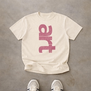 May include: A cream-coloured short-sleeve t-shirt featuring the word art printed vertically in a distressed, muted pink font across the centre of the chest. The garment has a standard crew neckline and a straight hemline, showcasing a simple design.