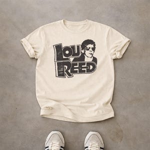 May include: A cream-coloured short-sleeve t-shirt featuring a black graphic print of Lou Reed wearing sunglasses. The design includes the text LOU REED in a bold, distressed font style. The shirt has a crew neckline and rolled sleeve cuffs.