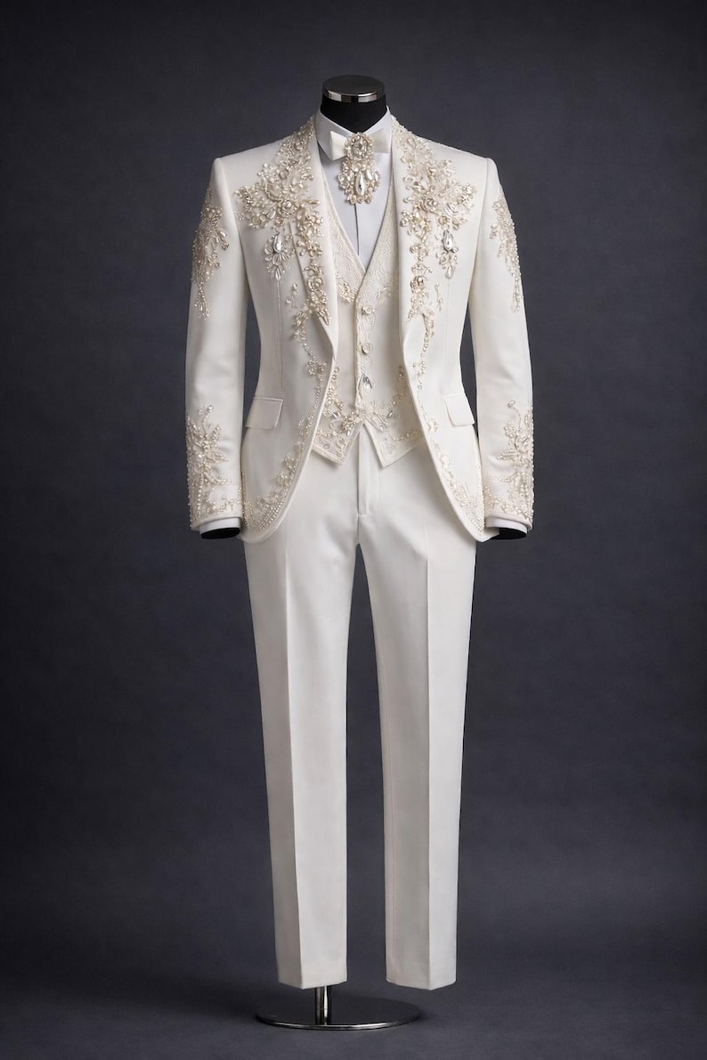 Op de afbeelding: Een formeel ivoorkleurig pak op een mannequin. Het pak bestaat uit een jasje, een vest en een broek. Het jasje en vest zijn versierd met ingewikkelde bloemenborduursels en sprankelende accenten. Het pak is getailleerd met een klassieke pasvorm.