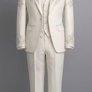 May include: Ivory-colored formal suit with intricate floral embroidery. The suit includes a jacket, vest, pants, and bow tie. The jacket and vest feature detailed embellishments. The suit is displayed on a mannequin against a gray backdrop.