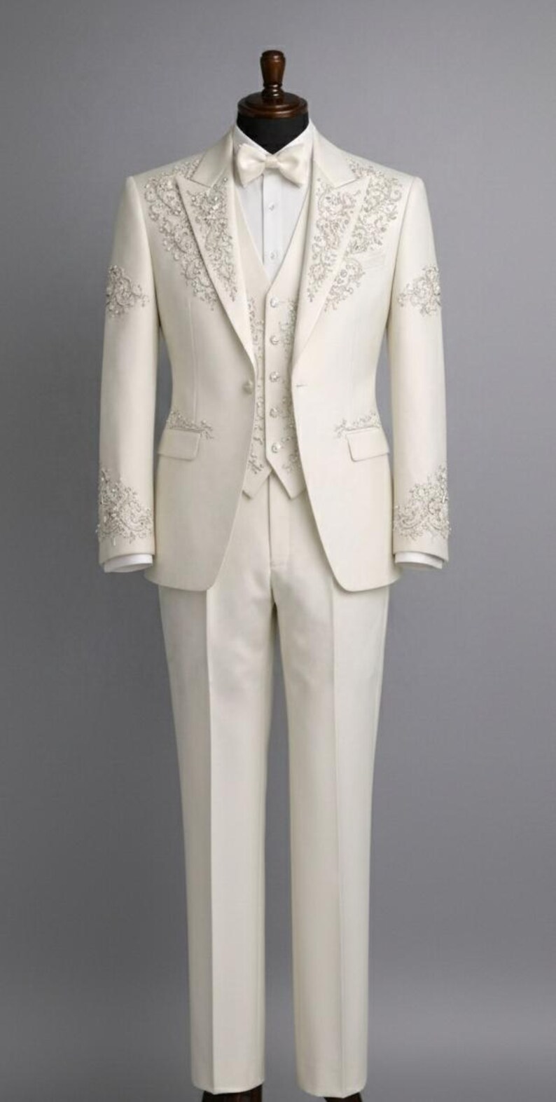 May include: Ivory-colored formal suit with intricate floral embroidery. The suit includes a jacket, vest, pants, and bow tie. The jacket and vest feature detailed embellishments. The suit is displayed on a mannequin against a gray backdrop.