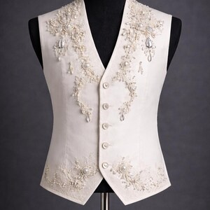 Op de afbeelding: Ivoor kleurige vest met ingewikkelde bloemenborduurwerk en versieringen. Het vest heeft een V-hals, knoopsluiting en is versierd met parels en glinsterende kristallen. Het ontwerp wordt op een mannequin getoond.
