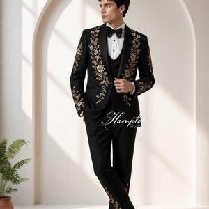 May include: A black tuxedo with gold floral embroidery. The suit includes a jacket, vest, and trousers. The jacket and trousers feature gold floral designs. The outfit is completed with a black bow tie and black dress shoes.