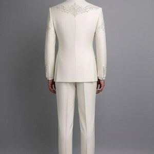 May include: Ivory-colored suit with intricate embroidered detailing on the back of the jacket and sleeves. The suit includes a tailored jacket and matching trousers. The ensemble is a formal wear item, suitable for special occasions.