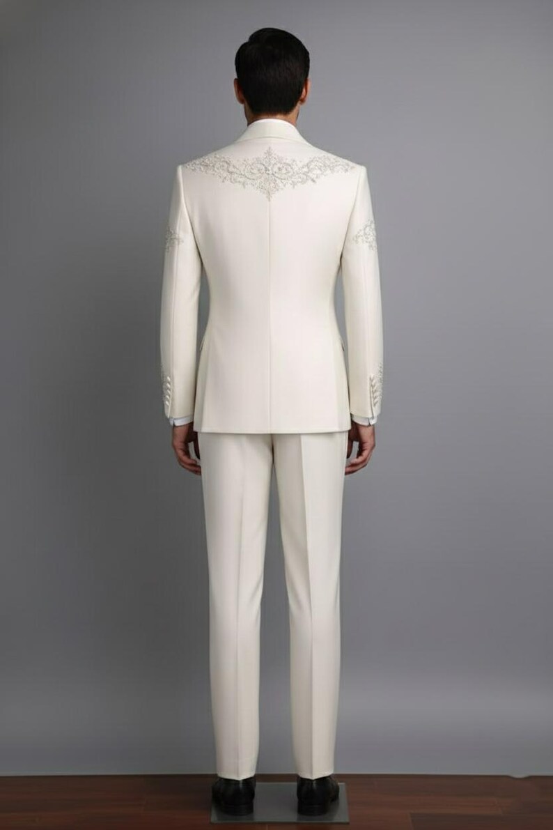 May include: Ivory-colored suit with intricate embroidered detailing on the back of the jacket and sleeves. The suit includes a tailored jacket and matching trousers. The ensemble is a formal wear item, suitable for special occasions.