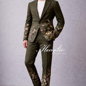 May include: Olive green suit with floral embroidery on the jacket sleeves and pant legs. The suit is paired with a white t-shirt and tan loafers. The text "Hampton Studio Inc." is visible on the pants.
