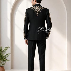 May include: Black suit with gold embroidery on the shoulders and sleeves. The suit includes a jacket and matching pants. The back of the jacket features an elaborate floral design. The suit is worn with a white collared shirt and black shoes. The text "Hampton Studios" is visible.