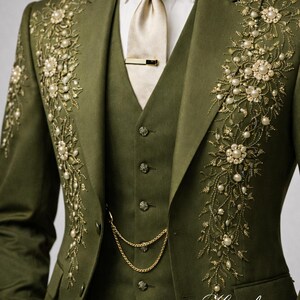 May include: Olive green three-piece suit with intricate floral embroidery and pearl accents. The suit includes a jacket, vest, and trousers, with a gold tie clip and chain. The suit is from Hampton Studio Inc.