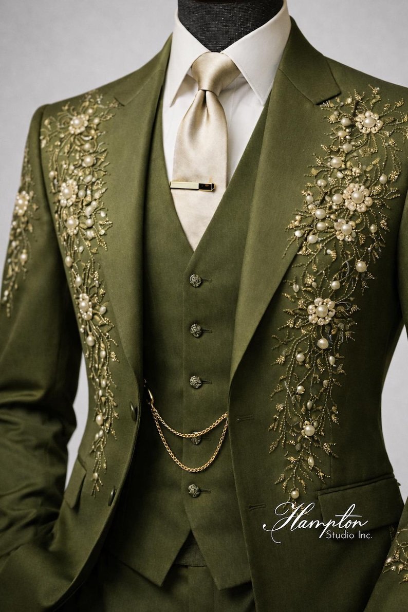 May include: Olive green three-piece suit with intricate floral embroidery and pearl accents. The suit includes a jacket, vest, and trousers, with a gold tie clip and chain. The suit is from Hampton Studio Inc.