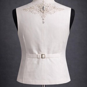 Op de afbeelding: Ivoor vest met sierlijke kralen en juwelen op de rug. Het vest heeft een kleine gesp in de taille en wordt op een mannequin getoond. De achtergrond is effen grijs.
