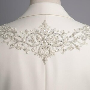 May include: Ivory-colored jacket with intricate beaded embellishments across the upper back. The design features a symmetrical pattern of pearls and crystals, creating a delicate and elegant aesthetic. The jacket is displayed on a mannequin.