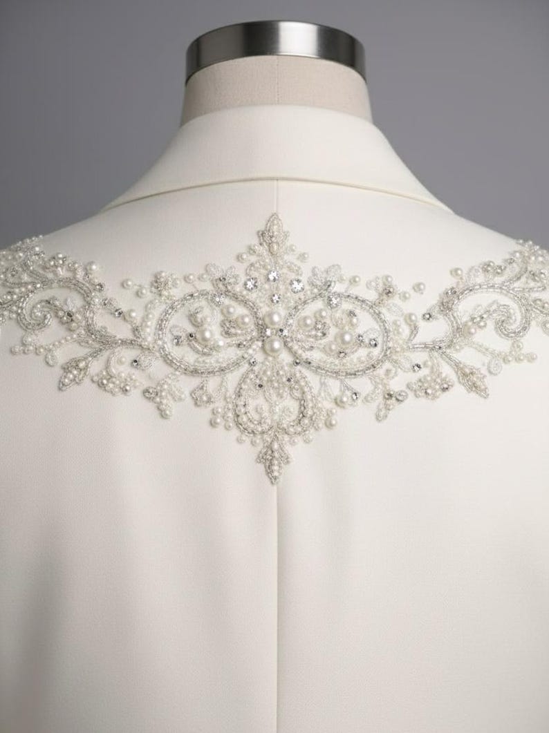 May include: Ivory-colored jacket with intricate beaded embellishments across the upper back. The design features a symmetrical pattern of pearls and crystals, creating a delicate and elegant aesthetic. The jacket is displayed on a mannequin.