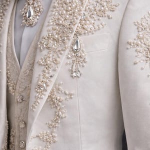 Op de afbeelding: Ivoor pak met ingewikkelde parel- en kristallen versieringen. De jas heeft bloemachtige ontwerpen en een decoratieve broche. Een bijpassende vlinderdas en vest maken het formele ensemble compleet, geschikt voor bruiloften of speciale evenementen.