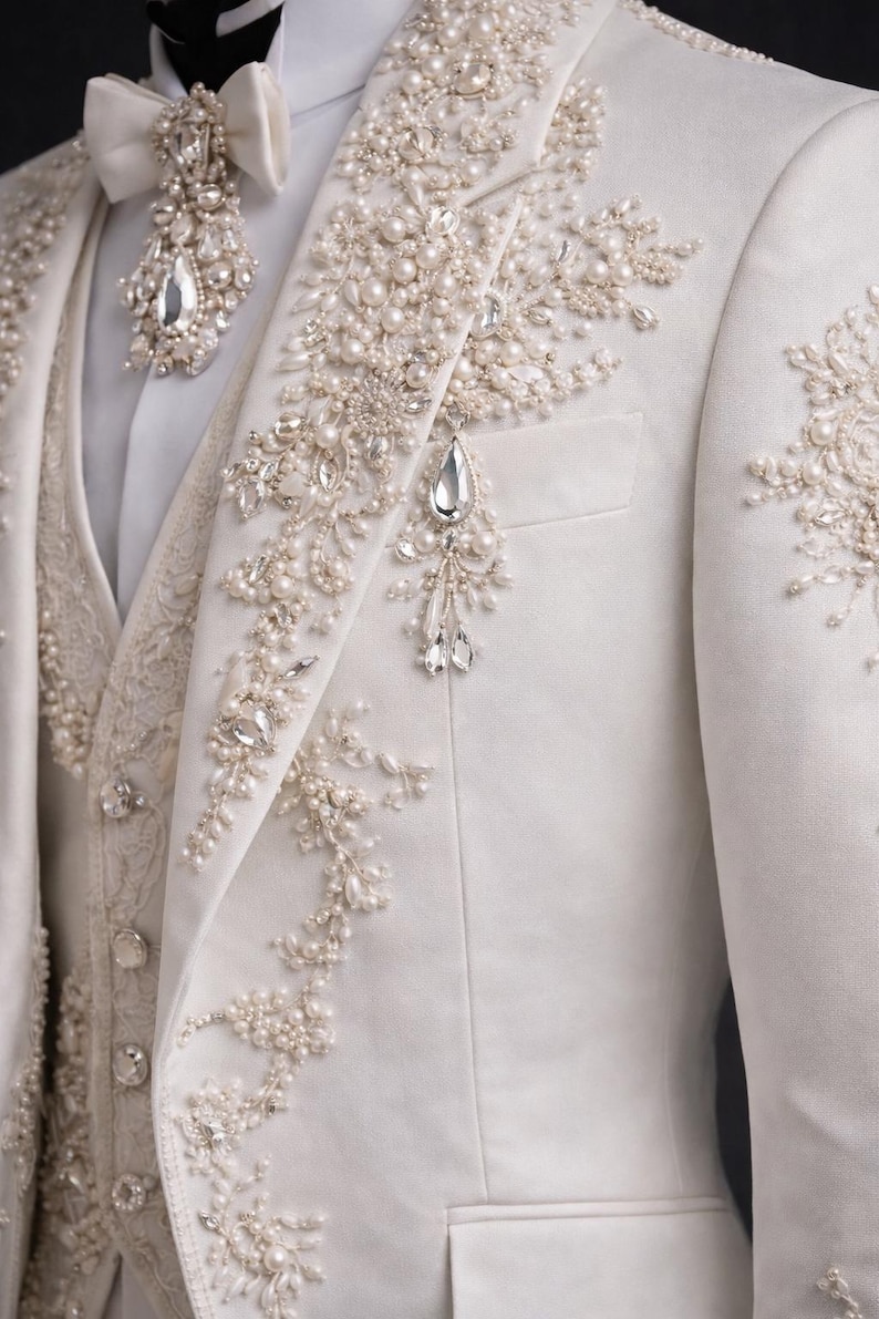 Op de afbeelding: Ivoor pak met ingewikkelde parel- en kristallen versieringen. De jas heeft bloemachtige ontwerpen en een decoratieve broche. Een bijpassende vlinderdas en vest maken het formele ensemble compleet, geschikt voor bruiloften of speciale evenementen.