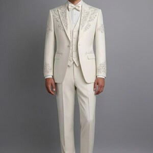 May include: Ivory three-piece suit with intricate floral embroidery on the lapels, sleeves, and vest. The suit includes a jacket, vest, and trousers, paired with a white dress shirt and bow tie. The ensemble is completed with white dress shoes.