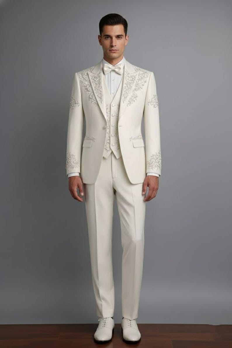 May include: Ivory three-piece suit with intricate floral embroidery on the lapels, sleeves, and vest. The suit includes a jacket, vest, and trousers, paired with a white dress shirt and bow tie. The ensemble is completed with white dress shoes.