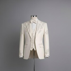 May include: Ivory three-piece suit with intricate floral embroidery. The suit includes a blazer, vest, and trousers, all in a solid ivory color. The blazer features a notched lapel and a bow tie. The suit is displayed on a mannequin against a gray background.